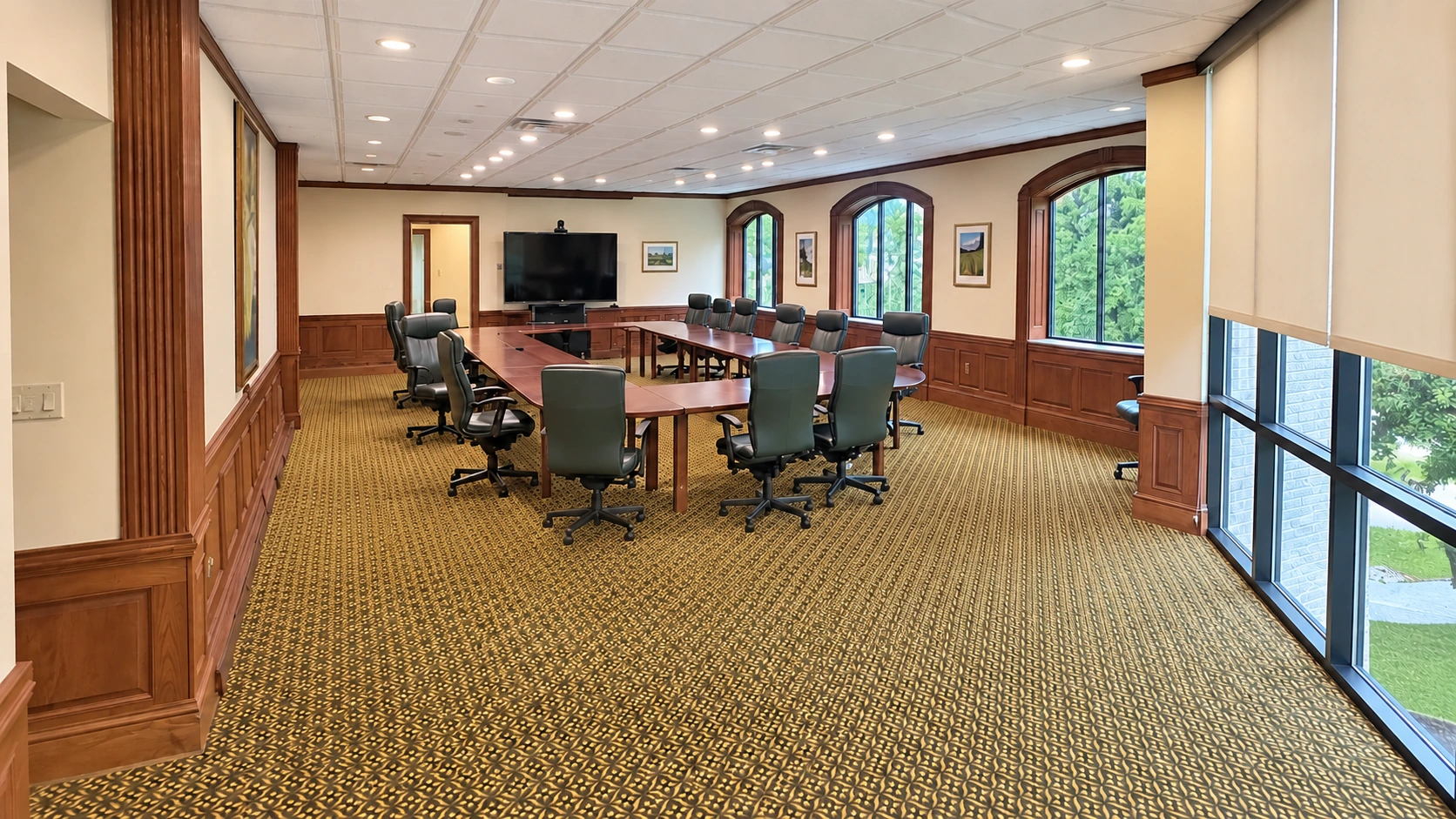 Wide view from the opposite end of the executive boardroom, showing conference seating, presentation screen and tall windows.