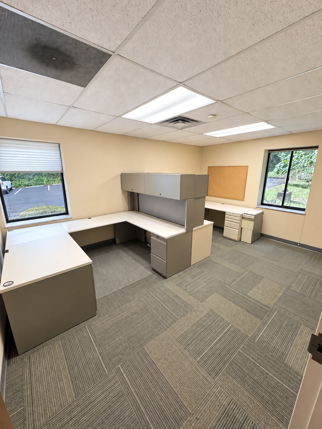 Office with two existing workstations, overhead storage and windows on two walls.
